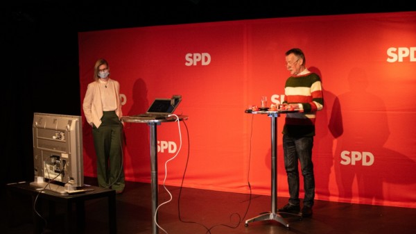 Zwei Personen stehen auf einer Bühne - hinter ihnen ein rotes SPD-Banner - vor ihnen ein Monitor und ein Tisch mit Laptop